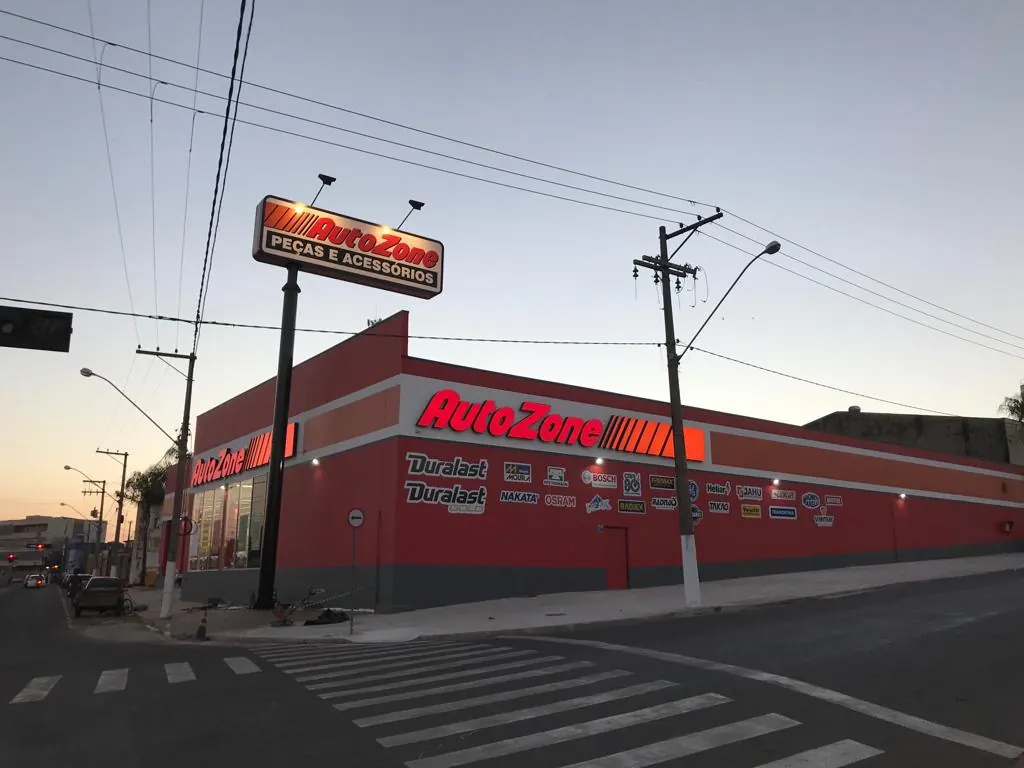 Fachada da loja Autozone em Leme, no fim da tarde, com letreiro iluminado, totem Peças e Acessórios e faixa de pedestres em primeiro plano