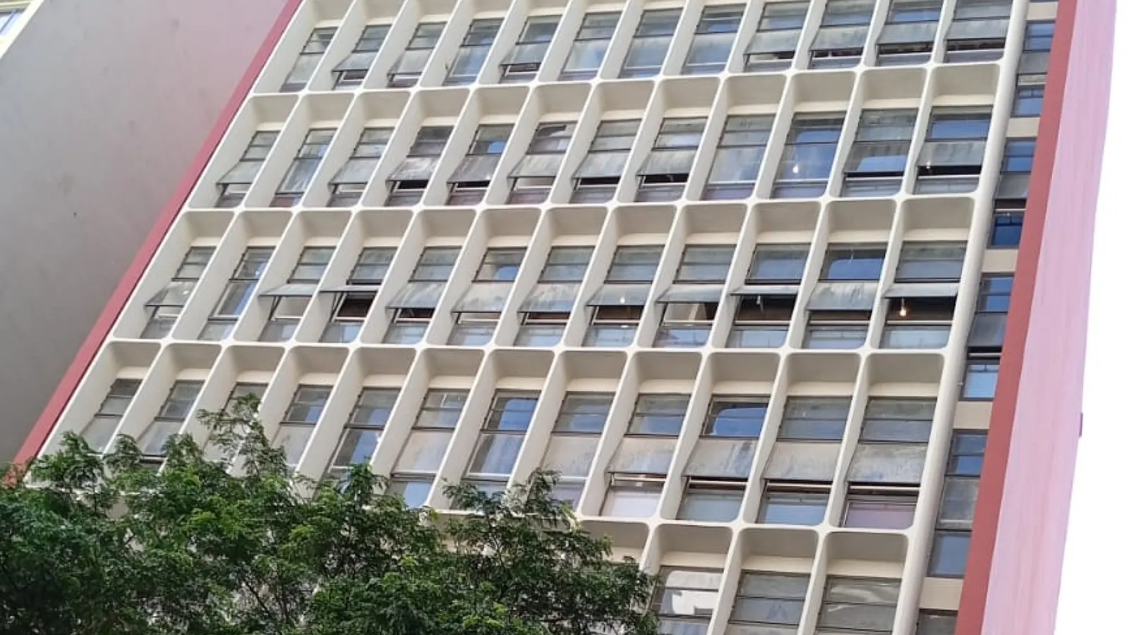 Fachada modernista do Edifício Dona Marcha com grelha de brises brancos e empenas vermelhas, vista de baixo entre árvores