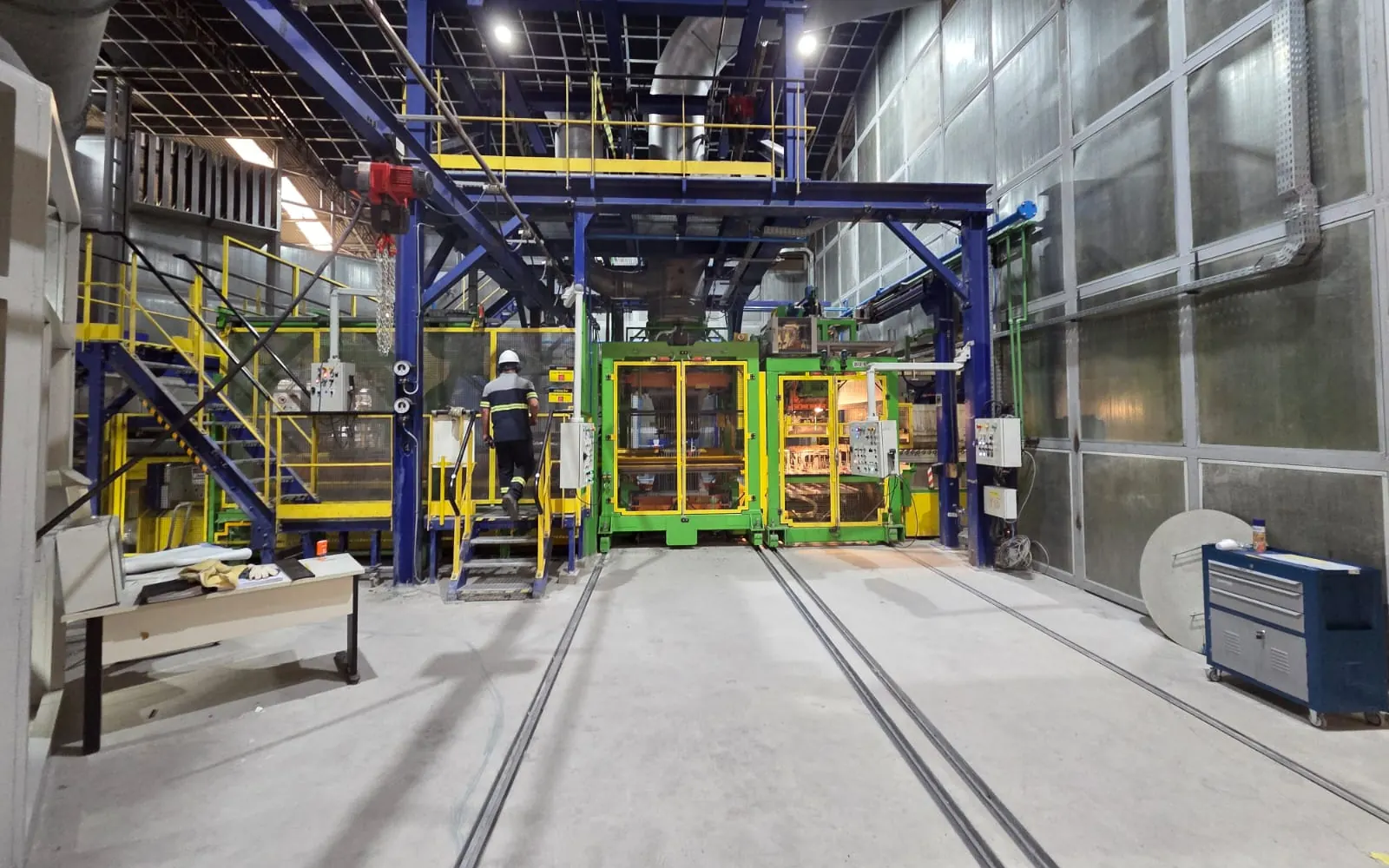 Linha de produção da fábrica Saint-Gobain em Mauá, com estrutura metálica azul, passarelas amarelas e trilhos embutidos no piso de concreto